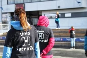 Foto: Person står med ryggen til med T-skjorte med teksten "Unio i streik". I bakgrunnen holder en person en appell.